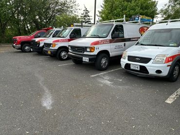 A line of commercial service vans parked in a lot with trees in the background.