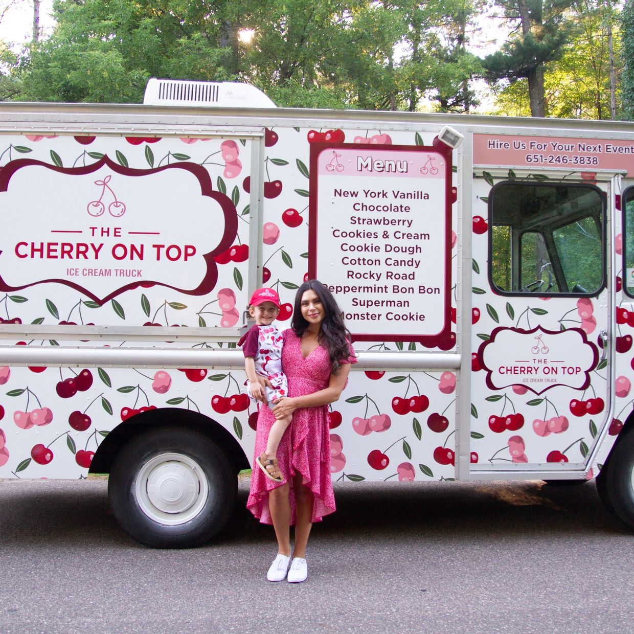 A woman in a pink dress holding a child stands in front of a cherry-themed ice cream truck.