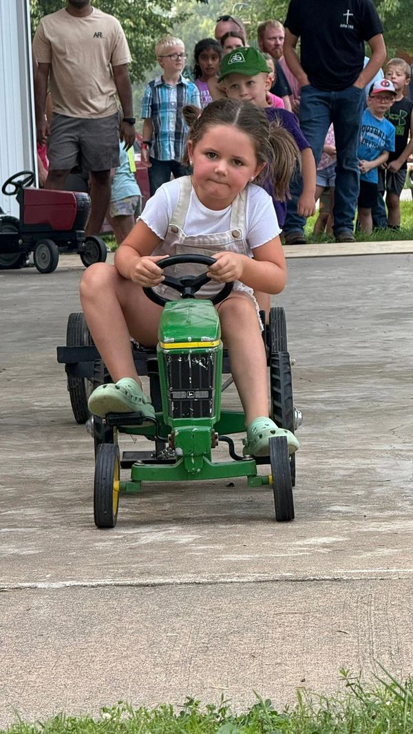 A determined girl rides a green toy tractor with people watching behind her.