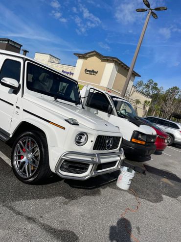 Luxury white Mercedes G-Class SUV parked beside other vehicles under a clear blue sky.