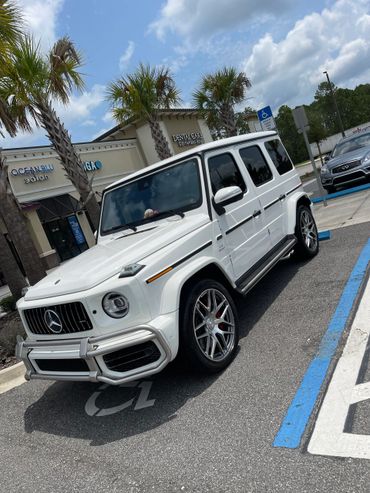 White Mercedes-Benz G-Class SUV parked in a handicap spot near palm trees and shops.