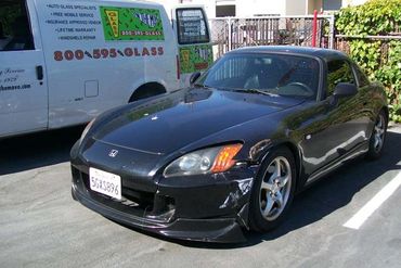 A black Honda S2000 sports car with front damage parked beside a service van, under bright sunlight.
