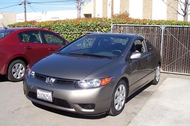 A silver Honda Civic coupe parked in a lot, with a red car in the background and green shrubs.
