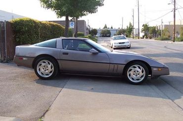 A gray Corvette C4 parked on the side of a road, with trees and buildings in the background