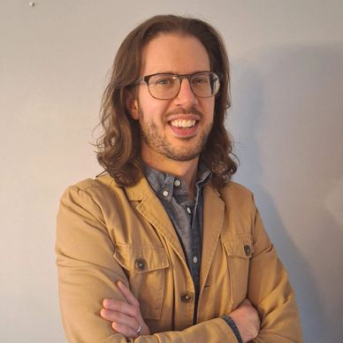 Man with glasses smiling, wearing a tan jacket and blue shirt, standing with arms crossed.