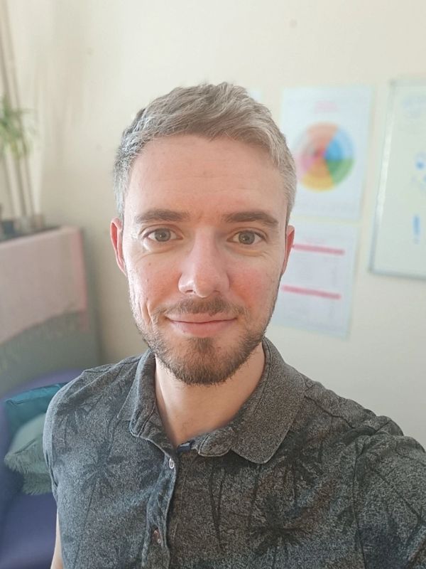 Smiling young man with short hair and beard in a casual shirt taking a selfie indoors.