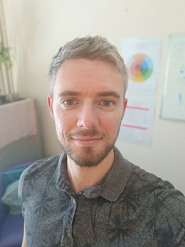 Smiling young man with short hair and beard in a casual shirt taking a selfie indoors.