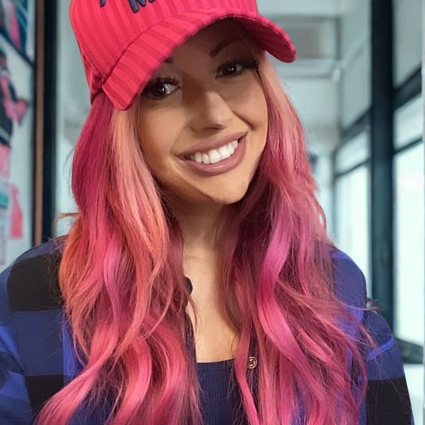 Smiling woman with long pink hair wearing a red cap and blue checkered shirt.