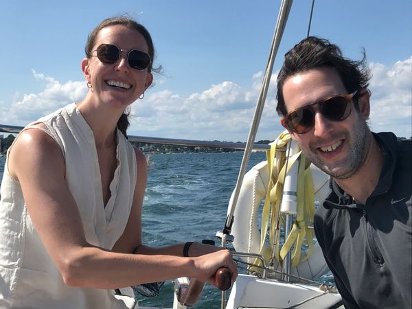 Two people smiling on a sailboat with sunny weather and blue sky.