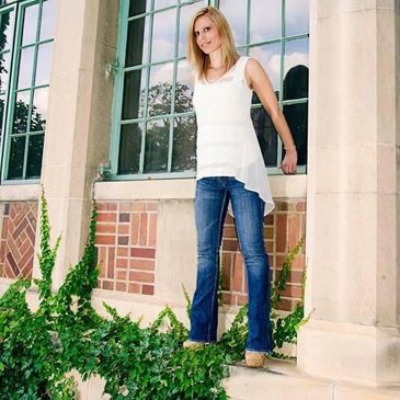 Woman in white top and jeans standing by a window.