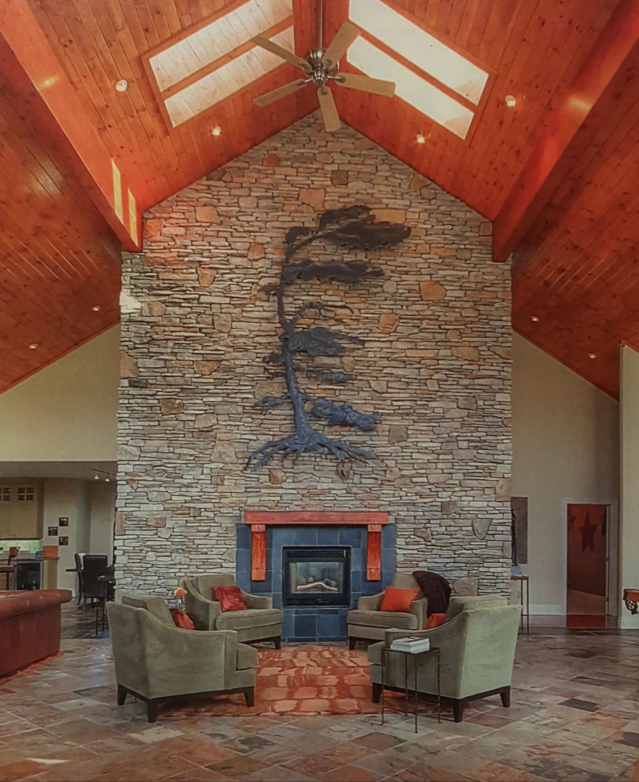 Finished metal tree sculpture on a stone wall above a fireplace. Sunlight streams through Skylights.