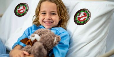 Smiling child in hospital bed with teddy bear and Ghostbusters logos.