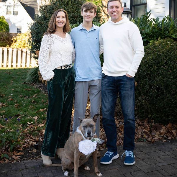 A family of three with their dog posing outdoors on a sunny day.