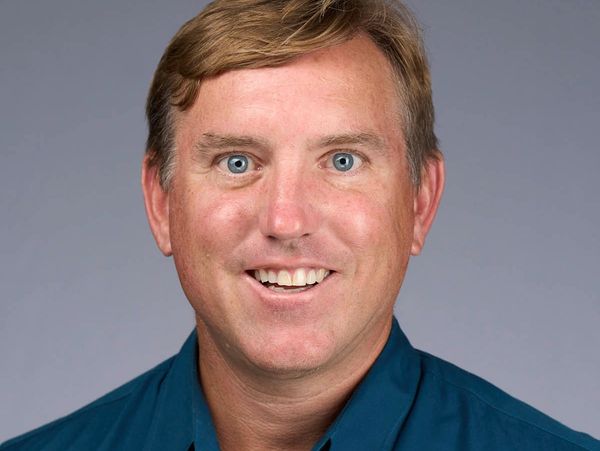 Smiling man with blue eyes in a teal shirt against a neutral background.