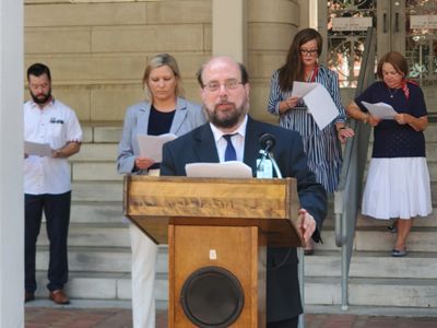 Man speaking at a podium with four people reading behind him on steps.