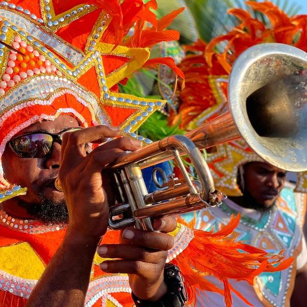 Two men dressed in full Junkanoo parade costumes with one of them playing a trumpet.