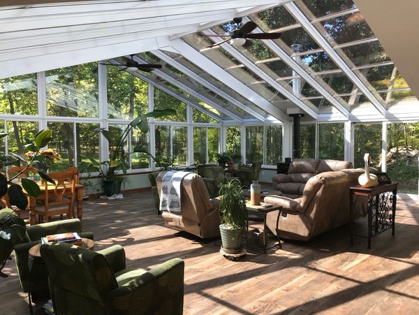 Spacious glass sunroom with sloped roof, cozy seating, plants, and natural woodland views