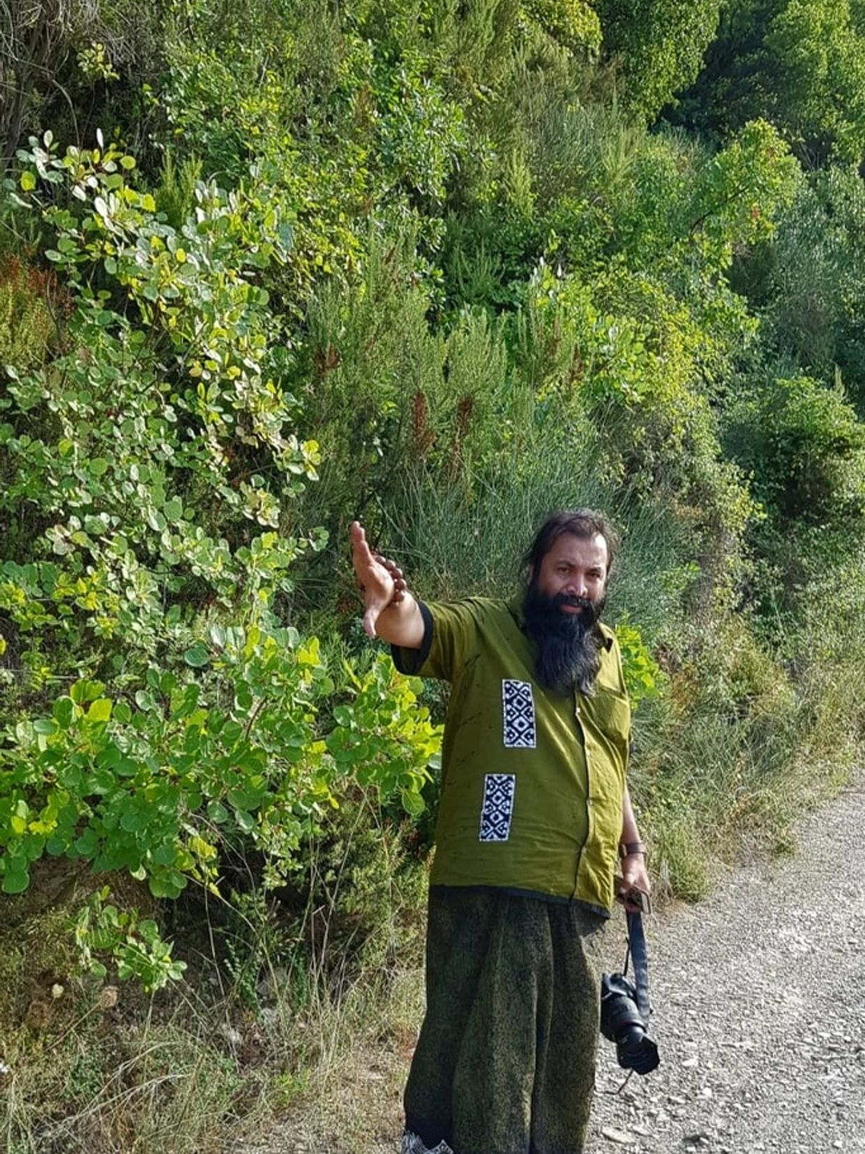 A bearded man in green waves beside a forest path holding a camera.