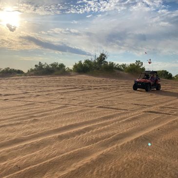 A dune buggy drives across sandy terrain under a partly cloudy sky at sunset.