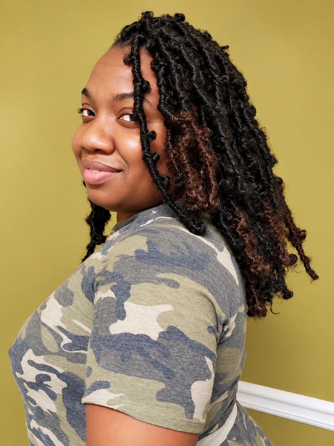Woman with curly hair in camo shirt against yellow background.