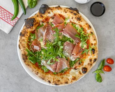 Wood-fired pizza topped with prosciutto and arugula on a white plate.