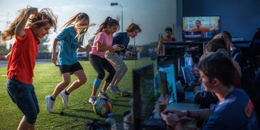 Children playing soccer outside and others gaming indoors on computers.