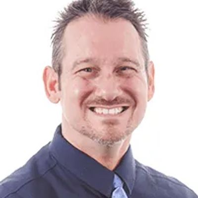 Smiling man in a navy shirt and light blue tie against a white background.