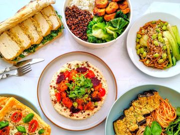 A colorful spread of plant-based dishes including sandwich, rice bowl, pasta, salad, and dip.