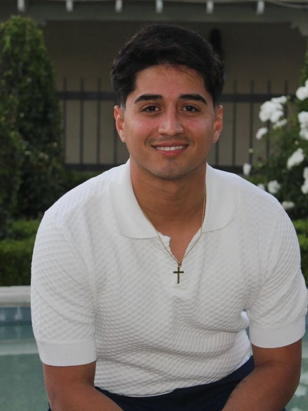 A young man in a white shirt with a cross necklace smiles outdoors.