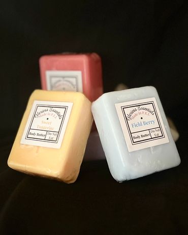 Three blocks of body butter in different scents and colors against a dark background.