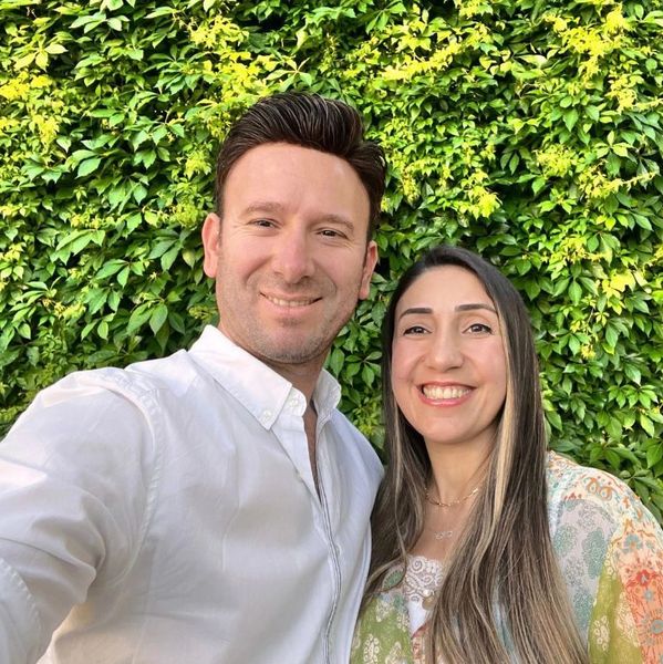 A smiling couple taking a selfie in front of lush green foliage.