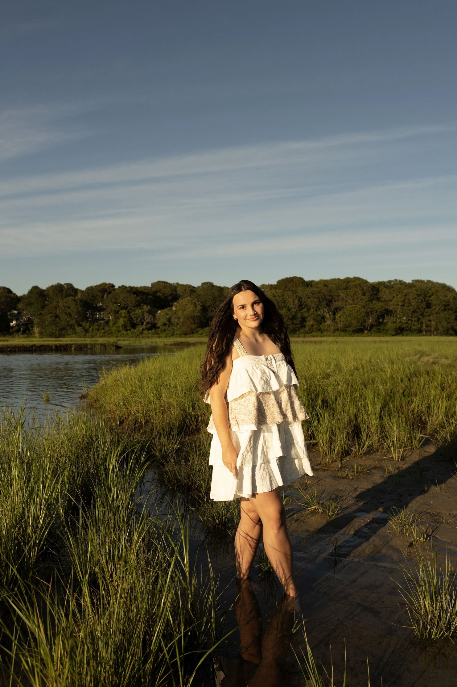 Young woman in a white dress standing in a marshy area with trees in the background.