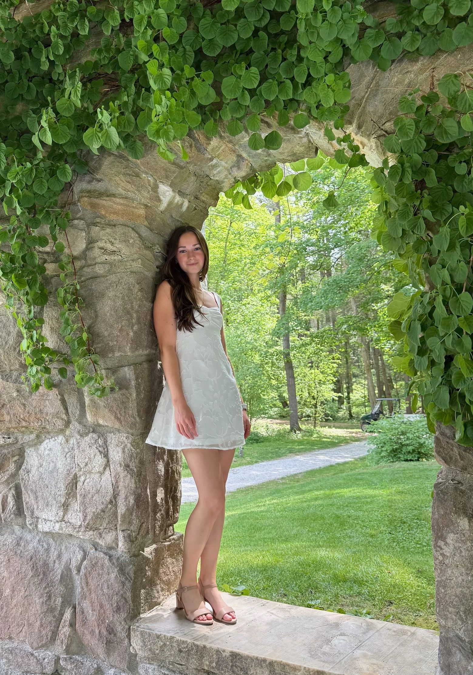 Young woman in a white dress stands under a stone arch covered with green ivy.