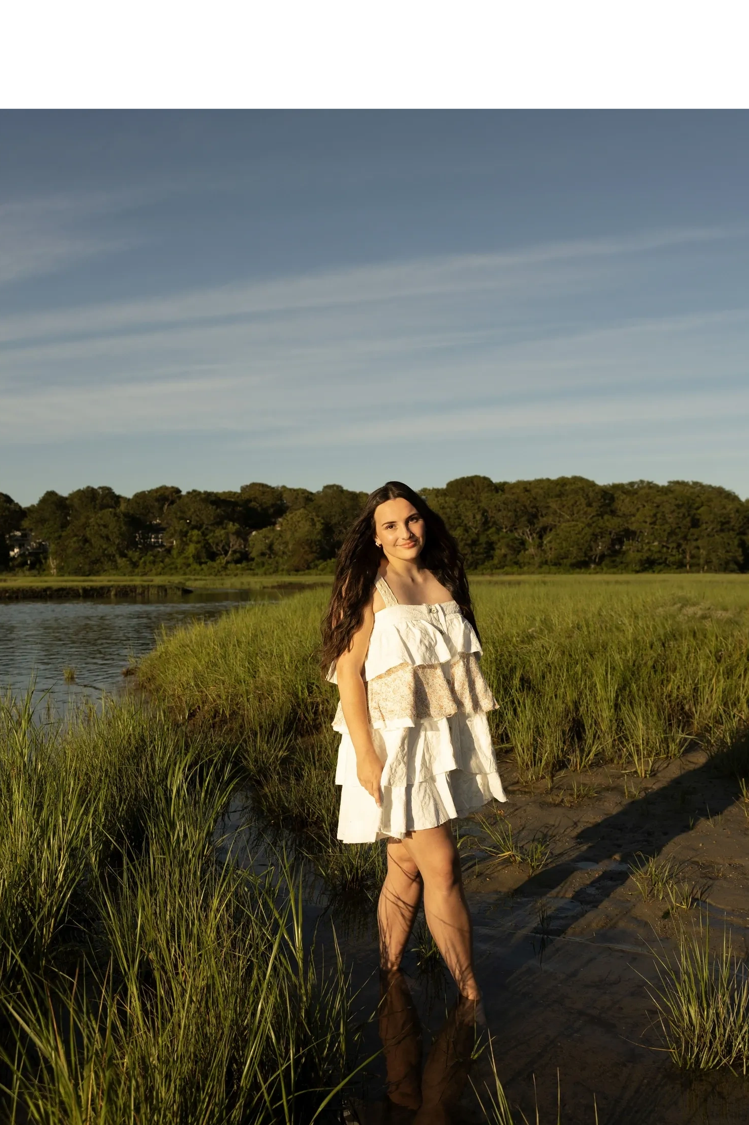 Young woman in a white dress standing in a marshy area with trees in the background.