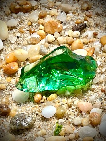 A vibrant green glass shard lies among small pebbles and sand.