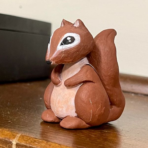 A small, hand-painted squirrel figurine with big eyes sits on a wooden surface.