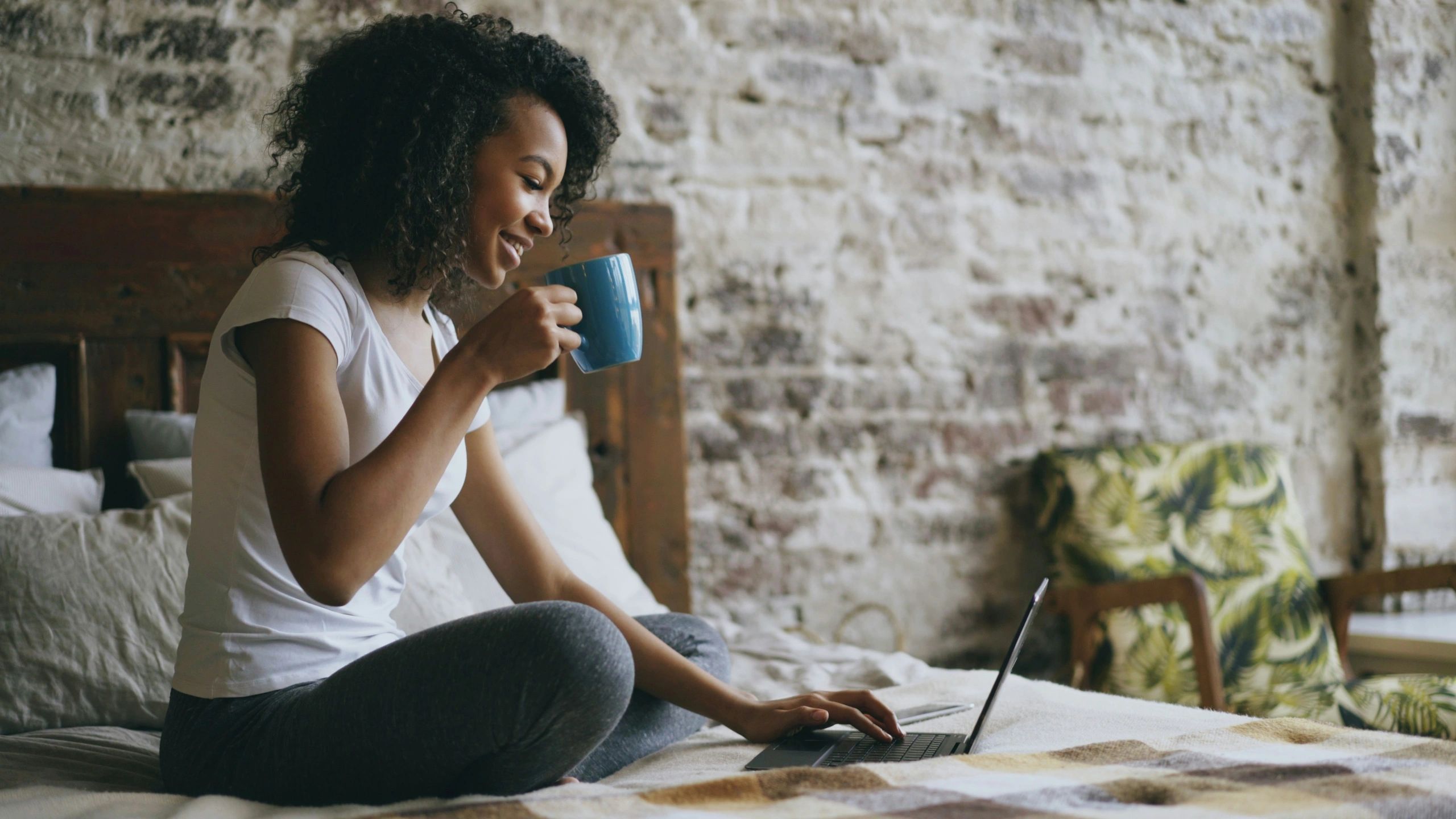 Woman sitting on her bed with a mug of coffee, smiling at her laptop