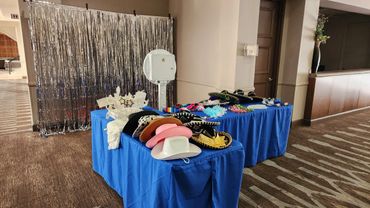 Photo booth setup with hats and props on blue-covered tables and a silver backdrop.