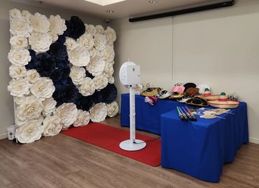 Photo booth setup with large flower backdrop, red carpet, and tables with props and hats.