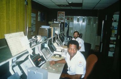 Two men working in a control room with multiple consoles.