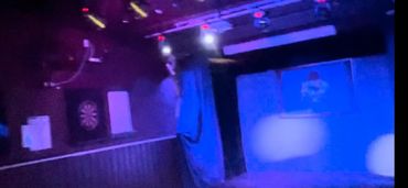 Dimly lit room with blue and purple lighting, dartboard on wall, and stage area.