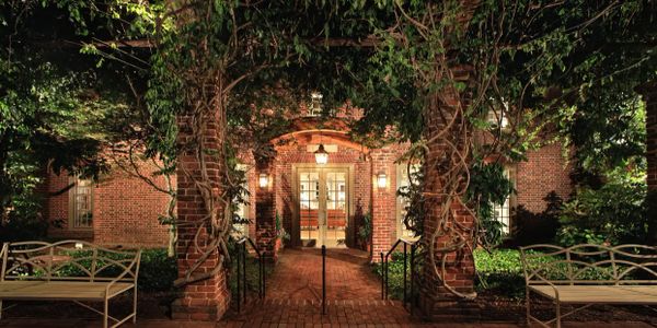 A warmly lit brick entrance framed by lush greenery and white benches.