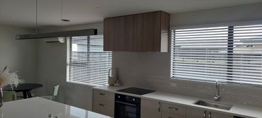 Modern kitchen with white countertops, wooden cabinets, and large windows with blinds.