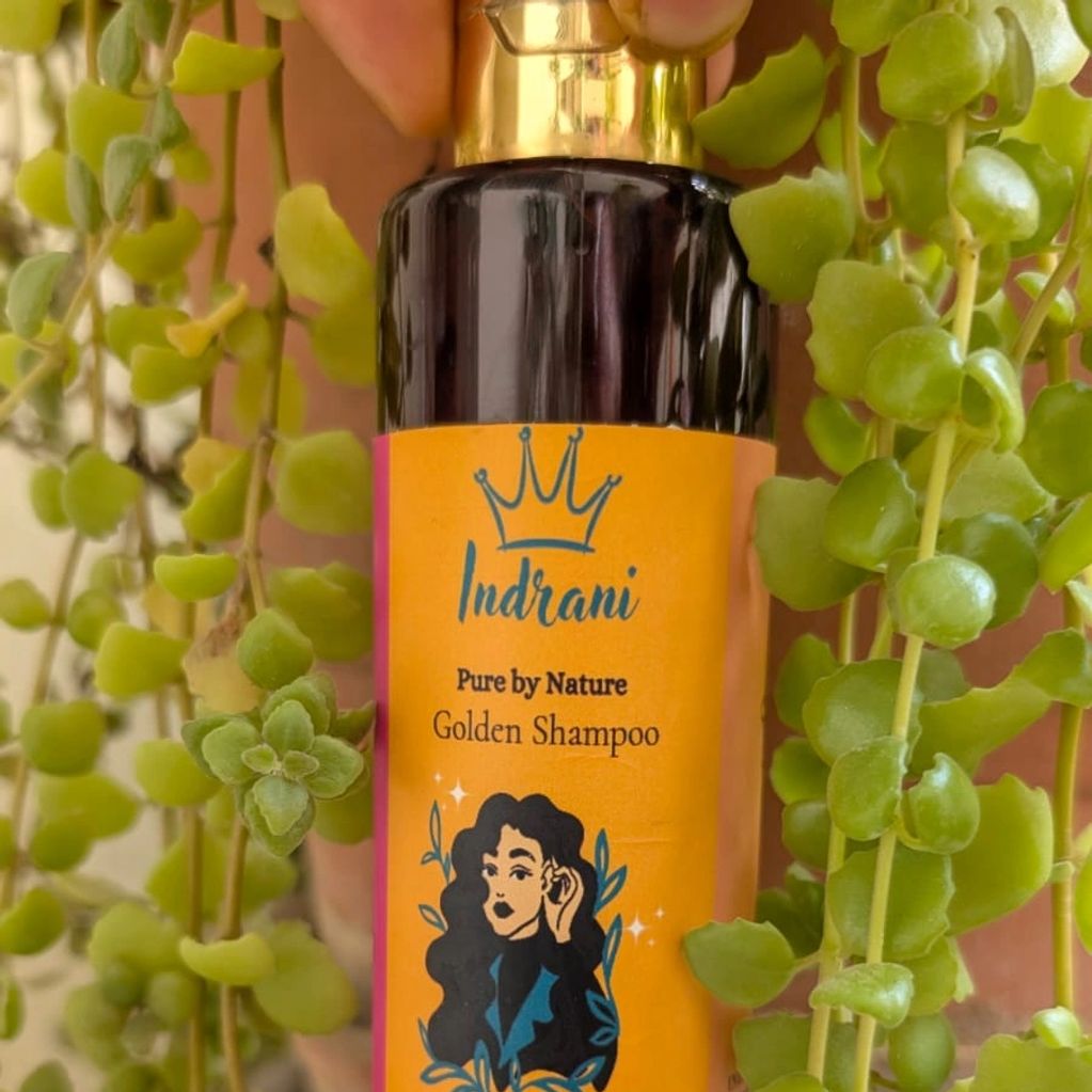 A hand holds Indrani Golden Shampoo amidst green plants.