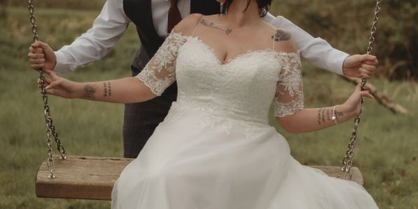 Bride in a white lace wedding dress on a swing held by groom.