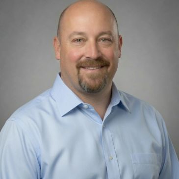 Smiling man in blue shirt against gray background.