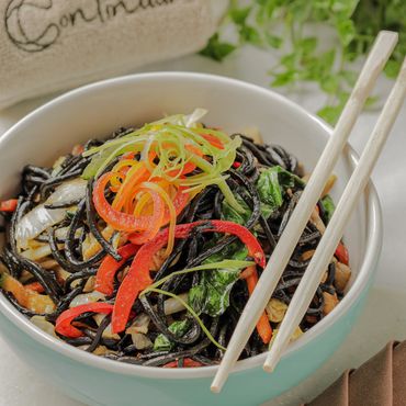 Bowl of black noodles with colorful vegetables and chopsticks.