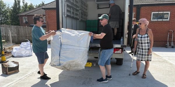 Two men lifting a large white sack outside a truck while a woman with a cane watches.