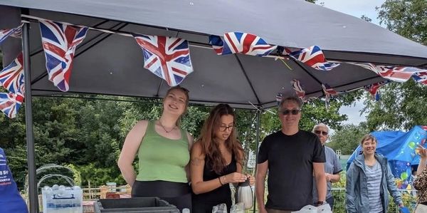 People gathered under a canopy with Union Jack flags, serving refreshments at a community event.