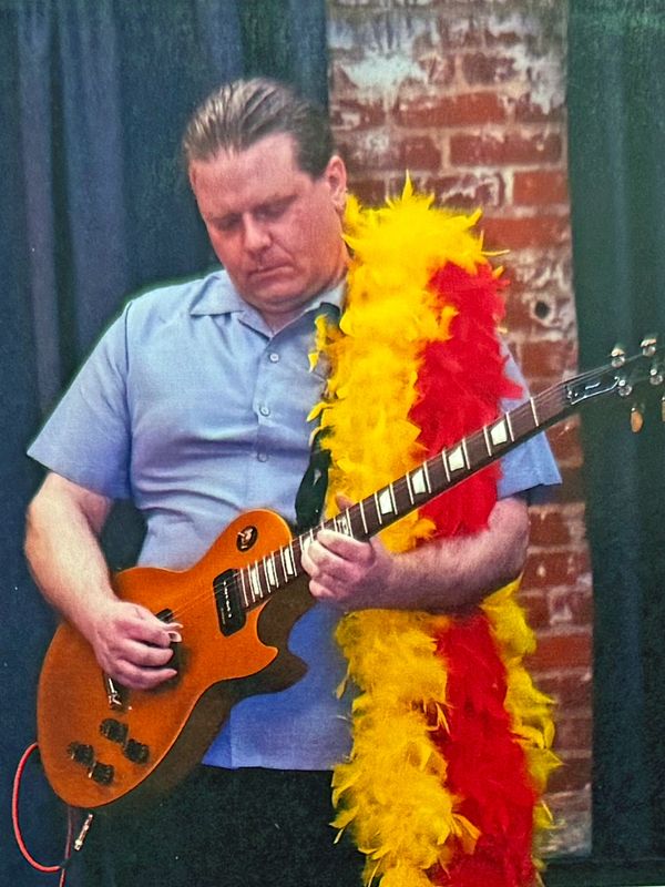 Man playing electric guitar with a colorful feather boa.